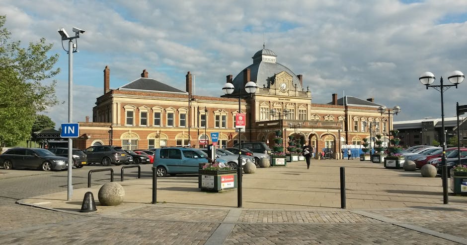 An exterior view of Finsbury Park station in London, showing a large historic brick building with a central clock and ornate architectural details. In front of the station, there is a paved pedestrian area with several parked cars, including a blue van and various other vehicles, arranged along the perimeter. The scene includes black metal bollards and a few large spherical stone barriers on the pavement, which is surrounded by street lamps and flower planters. A person dressed in dark clothing is walking across the open space, while others are near the station entrance. The sky is partly cloudy, with sunlight illuminating the scene, suggesting daytime. This image captures an active urban environment typical of borough relocations involving professional removals companies such as Finsbury Park Removals, who handle home relocation, furniture transport, packing, and moving logistics near Finsbury Park Station.