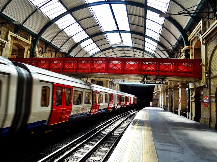 An exterior view of Finsbury Park station in London, showing a large historic brick building with a central clock and ornate architectural details. In front of the station, there is a paved pedestrian area with several parked cars, including a blue van and various other vehicles, arranged along the perimeter. The scene includes black metal bollards and a few large spherical stone barriers on the pavement, which is surrounded by street lamps and flower planters. A person dressed in dark clothing is walking across the open space, while others are near the station entrance. The sky is partly cloudy, with sunlight illuminating the scene, suggesting daytime. This image captures an active urban environment typical of borough relocations involving professional removals companies such as Finsbury Park Removals, who handle home relocation, furniture transport, packing, and moving logistics near Finsbury Park Station.