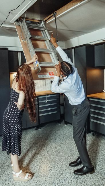Two individuals are engaged in a home relocation process inside a modern kitchen. The person on the right, a man dressed in a light blue shirt and dark trousers, is lifting and guiding a wooden ladder attached to a metal frame that extends from the ceiling, likely for access to an attic or high storage area. The woman on the left, wearing a navy blue dress with white polka dots and white sandals, is assisting by holding the ladder steady with both hands. The kitchen features black cabinetry with silver handles, a wooden work surface, and a pegboard on the wall with various tools and supplies visible beneath the ladder. The scene is illuminated by ceiling and ambient lighting, emphasizing the careful handling of furniture or equipment during the packing and moving process. This image illustrates professional removals services offered by Finsbury Park Removals, specializing in furniture transport and home relocations, ensuring safe and efficient logistics for house moves.