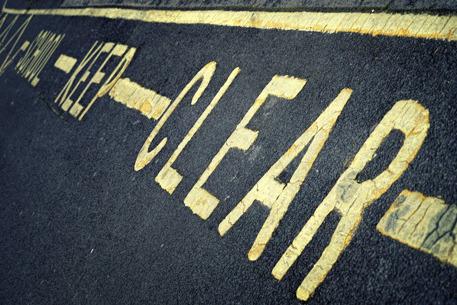 Close-up view of a yellow 'CLEARWAY NO STOPPING' road marking on dark asphalt pavement outside a property, indicating a restriction zone for vehicles. The painted text is slightly cracked and weathered, running diagonally across the image, with a textured surface showing tire marks and small debris nearby. Behind the pavement, there is a street curb and part of a building entrance, suggesting the location is adjacent to a residential or commercial property in Green Lanes, Finsbury Park. This scene illustrates the importance of understanding local parking and loading regulations when planning a home relocation or furniture transport, often coordinated by specialists such as Finsbury Park Removals, to ensure smooth loading and unloading within legal bounds.