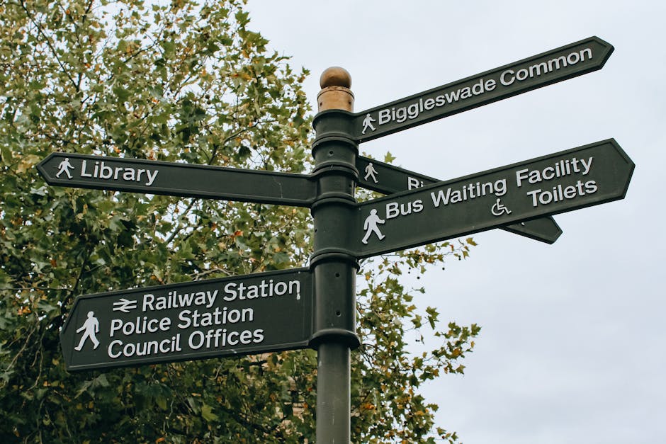 A black multi-directional street signpost with five signs mounted on a central pole, situated outdoors against a background of green leafy trees and an overcast sky. The signs indicate directions to Biggleswade Common, Library, Railway Station, Police Station, Council Offices, Bus Waiting Facility, and Toilets, with some including icons for pedestrians and accessibility. The signpost is part of an urban or residential area, and its positioning suggests navigation guidance for local landmarks and public facilities. This image relates to house removals and relocation services, as shown on the Finsbury Park Removals' N4 removals guide page, reflecting the importance of clear signage when organizing home or furniture transport in residential locations.