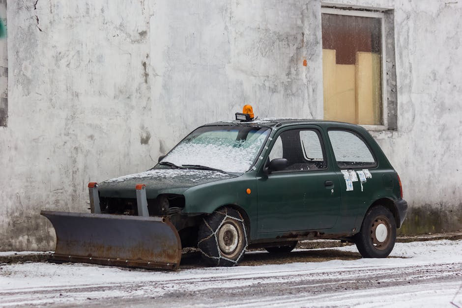 A small, green, compact vehicle parked outside a grey concrete building during winter, with snow covering the ground and parts of the car. The vehicle has a snow-covered roof, and a snow-dusted front windshield, with a roof-mounted warning light visible. The car's front bumper and headlights are missing, exposing internal components, and the front left tire is replaced with a snow chain attached to the wheel, indicating a need for enhanced traction in icy conditions. The vehicle is positioned close to a window with wooden frames, and nearby, there are cardboard or paper notices taped to the rear side window. This scene, captured during a cold, snowy day, may depict a vehicle awaiting repair or disposal, reflecting winter weather challenges. For house removals or transport services, Finsbury Park Removals can assist with vehicle logistics or move-related transportation.